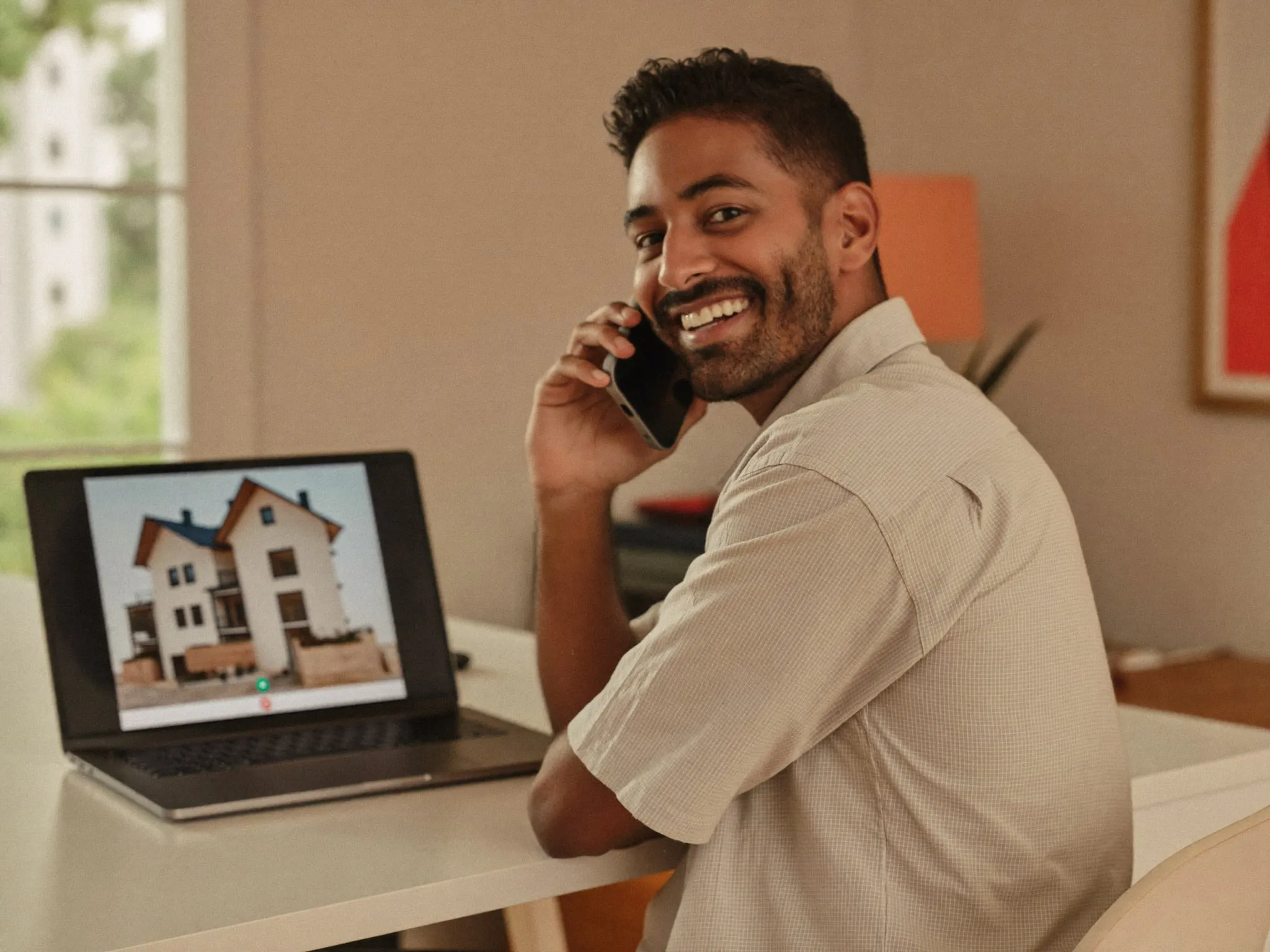 Illustrative: smiling NRI client on a phone call, with his home construction progress on the laptop screen